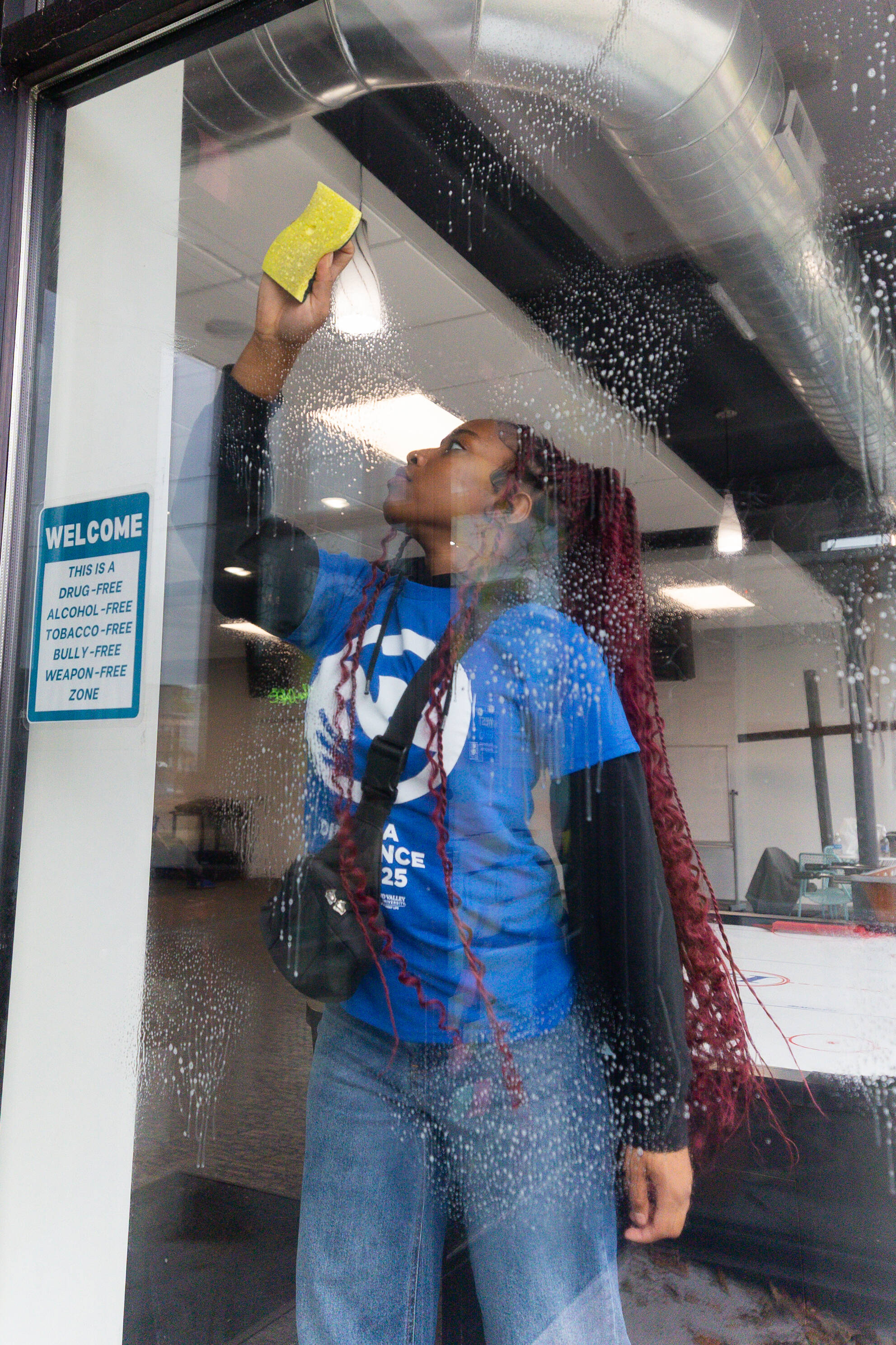 A person in a blue Make a Difference Day shirt cleans the inside of a glass window with a sponge.
